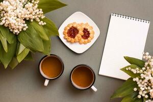 Two mugs with tea, cookies, a notebook and bouquets of lilies of the valley on a green background. Pause for rest, slow life, planning, goal setting. Top view photo