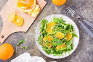 Diet vegetarian vitamin salad of orange slices and a mix of arugula, chard and mizun leaves on a plate and a cutting board with peeled orange on the table. Top view photo