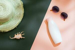 Beach vacation. A straw hat, sunglasses, a bottle of sunscreen cream and shells on a green-pink cardboard in the shadows of a tree leaves. Items for sunbathing and tanning. Top view photo