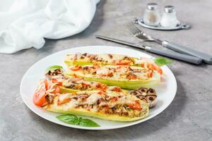 Ready-to-eat baked zucchini halves stuffed with cheese and tomato and basil leaves on a plate on the table. Vegetable dishes, healthy food photo