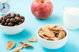 bochas con cereal y chocolate pelotas, un vaso de leche, un manzana y un alarma reloj en un azul antecedentes. programado desayuno. de cerca foto