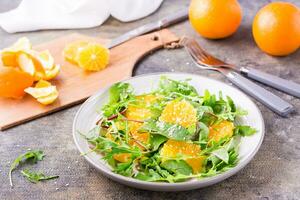 dieta vegetariano vitamina ensalada de naranja rebanadas y un mezcla de Rúcula, acelga y mizun hojas en un plato y un corte tablero con pelado naranja en el mesa foto