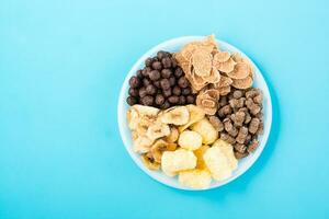 A plate with different types of breakfasts and snacks oatmeal, cereal, chocolate balls, banana chips and rye bran on a blue background. Top view photo