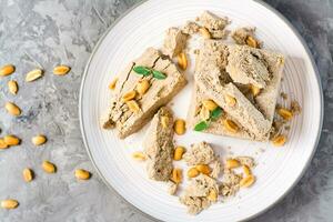 Pieces of sunflower and peanut halva and mint leaves on a plate and nuts nearby on the table. Caloric oriental delicacy. Top view. Close-up photo