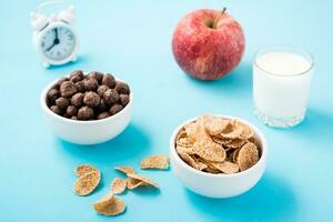 bochas con cereal y chocolate pelotas, un vaso de leche, un manzana y un alarma reloj en un azul antecedentes. programado desayuno foto