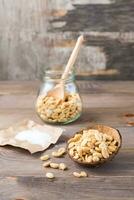 Salted peanuts in a bowl and in a jar with a spoon and salt on paper on a wooden table. Healthy vegetarian food. Rustic style. Vertical view photo