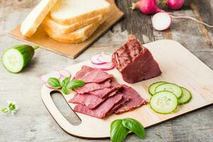 Fresh sliced marbled beef pastrami, cucumber, radish and basil on a cutting board. American delicacy. Rustic style photo