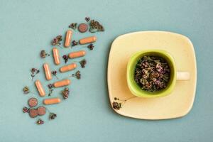 Medicinal capsules, pills and herbs in the form of a branch on a green background and a cup with oregano. Alternative medicine. Top view photo