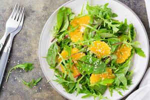 Diet vegetarian vitamin salad of orange pieces and mix of arugula, chard and mizun leaves on a plate on the table. Top view. Close-up photo