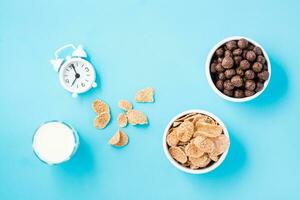 bochas con cereal copos y chocolate pelotas, un vaso de Leche y un alarma reloj en un azul antecedentes. programado desayuno, elección de platos. parte superior ver foto