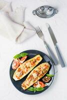 Ready-to-eat baked zucchini halves filled with cheese and tomato and basil leaves and cutlery on a black plate on a white table. Vegetable menu, healthy food. Top and vertical view photo