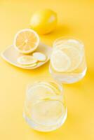 Sparkling water with lemon and ice in glasses and lemon slices on a saucer on a yellow background. Detox drink. Vertical view photo