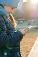 un niña en un chaqueta es participación un teléfono inteligente y mensajes de texto un mensaje en el rayos de el noche Dom. Internet surf foto