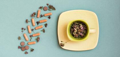 Medicinal capsules, pills and herbs in the form of a branch on a green background and a cup with oregano. Alternative medicine. Top view. Web banner photo