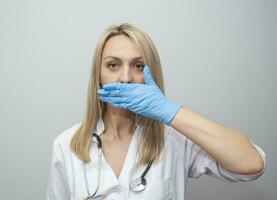 Confused young female doctor covering mouth isolated on gray background. medicinal secret. photo