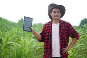 Handsome Asian man farmer wear hat, red plaid shirt, hold smart tabet, pose hand on waist, stand at maize garden. Concept. Agriculture occupation, survey and research to develope crops photo