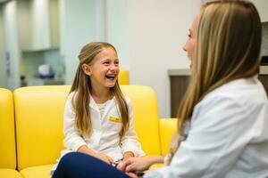 un dentista en un blanco Saco negociaciones acerca de el dientes a un niña en un amarillo vestir quien mentiras en el sofá foto