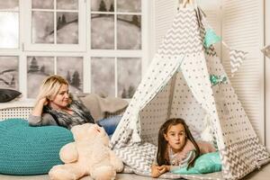 un joven madre en un gris suéter y hija son sentado en el para niños habitación en invierno. el Navidad y nuevo años interior. día festivo. familia concepto. foto