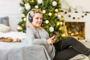 contento mujer disfrutando su música a Navidad relajante en un sofá en frente de el árbol con un radiante sonrisa abrochando su móvil a su pecho. foto