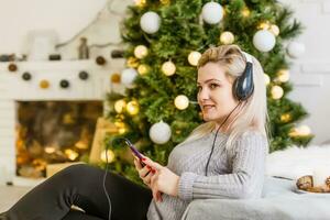 contento mujer disfrutando su música a Navidad relajante en un sofá en frente de el árbol con un radiante sonrisa abrochando su móvil a su pecho. foto