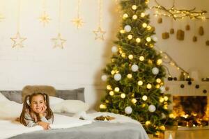 muy bonito encantador pequeño niña acostado en el cama y mirando a el imagen en el antecedentes de sonriente Navidad arboles en brillante interior de el casa foto