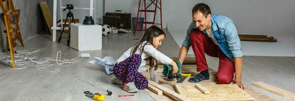 Side view of young happy father with his little daughter who do repair at home, happy daughter paints arms of father with help of platen photo