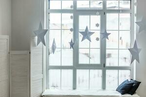 Large bright window. On the window-sill is a gray knitted plaid with gifts and sparkles with herlands. Paper Christmas stars are suspended on the background of the window. photo