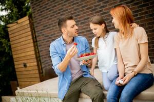 Family with a mother, father and daughter sitting outside on steps of a front porch of a brick house and eating strawberries photo