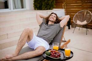 Young man relaxing on deck chair with fresh fruits and cold orange juice by the swimming pool in the house backyard photo