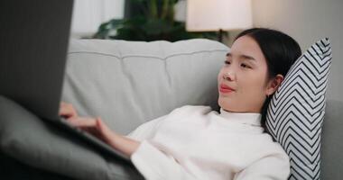 metraggio di contento giovane asiatico donna utilizzando un' il computer portatile computer mentre dire bugie su il divano nel il vivente camera. benessere a casa, rilassante e stile di vita concetti. video