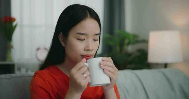 metraggio di contento giovane asiatico donna potabile un' caffè mentre seduta su il divano nel il vivente camera. benessere a casa, rilassante e stile di vita concetti. video