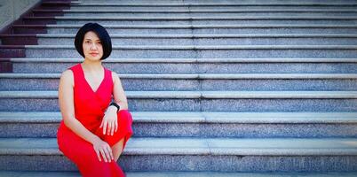 Beautiful or pretty Asian woman with short hair wearing red dress sitting on marble stair, step or staircase with copy space. Portrait fashion, casual style, smiling and relaxing. photo