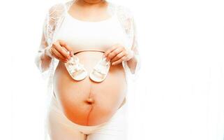 Closeup pregnant belly with cute tiny baby shoes. Concept of pregnancy, gynecology, preparation. Beautiful pregnant woman in shirt in white background. Woman holding big pregnant stomach. photo