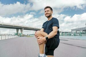 A confident male athlete training does warm-up exercises. Jogging in the afternoon in the city. Runner trainer in fitness clothes T-shirt. Uses a fitness watch. photo