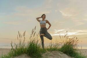 Female Fitness Trainer on the sand beach sunset background.  Meditation and harmony and balance of a person, mental health.   Practicing yoga exercises asana. photo
