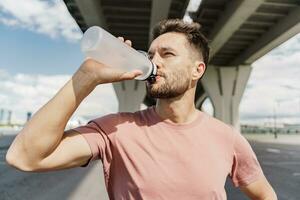 A sports man drinks water from a sports bottle, an athlete who is training does warm-up exercises. Jogging in the afternoon in the city. Runner trainer in fitness clothes T-shirt. photo