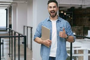 Happy young man standing in the office with thumb up, holding laptop, looking at camera. Copy space. Startup, coworking, management concept photo