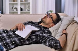 Bookish tranquility. A young man with facial hair wearing glasses fell asleep while reading a book on a plush couch in the inviting ambiance of his living room. Relaxation and literature concept. photo