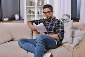 Cheerful bearded man in glasses immersed in a captivating book, savoring a serene moment in his cozy living room. Relaxation and literature concept for peaceful minds. photo