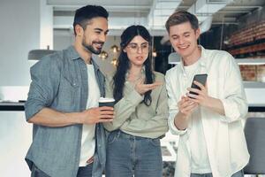 Young colleagues standing together and looking at phone screen chatting during coffee break. Startup, coworking, management concept photo