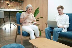 Smiling man using laptop, adult woman holding smartphone. Two entrepreneurs sitting in armchair in the office. Startup, coworking, management concept photo