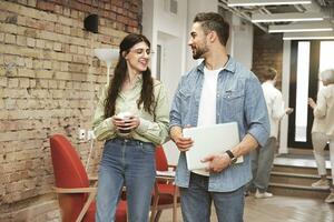 Happy young man with laptop and woman with coffe talking while walking along office corridor. Business, coworking, management concept photo