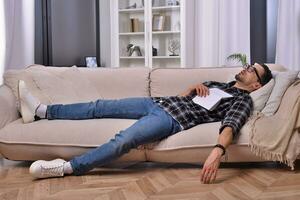 Bookworm's slumber. A man, donning stylish glasses, drifted off to sleep while indulging in a book on a comfortable couch in his inviting living room. Peaceful dreams amid the world of literature. photo