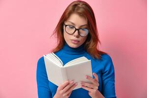 Focused redhead young pretty lady with glasses reading a book with an empty white cover isolated on a pink colored background. Book lovers and education concepts. photo