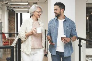 Smiling adult woman and young man walking in the office while talking, holding coffee. Startup, coworking, management concept photo