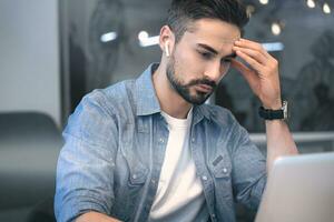 Young businessman sitting at desk and surfing net on laptop at office while using wireless headphones. Startup, coworking, management concept photo