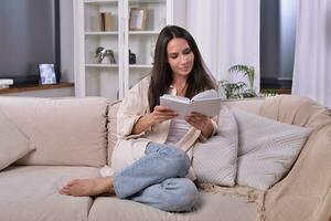 Serene woman engrossed in a book on her cozy living room sofa. The soft ambiance and peaceful expression reveal her complete immersion in the captivating world of literature. Tranquil reading. photo