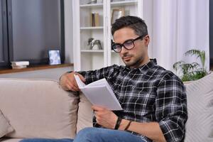 un intelectual joven hombre, poniéndose elegante anteojos, sumerge él mismo en un cautivador libro. con acogedor decoración, él disfruta un tranquilo leyendo sesión en el sofá en su encantador hogar. foto