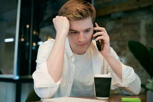 Young man sitting at his workplace in the office while talking on mobile phone, coffee on the table. Startup, coworking, management concept photo