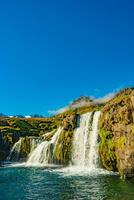Wonderful waterfall named Kirkjufellsfoss with a Kirkjufell church like iconic mount in Western Iceland, at blue sky and sunny day photo
